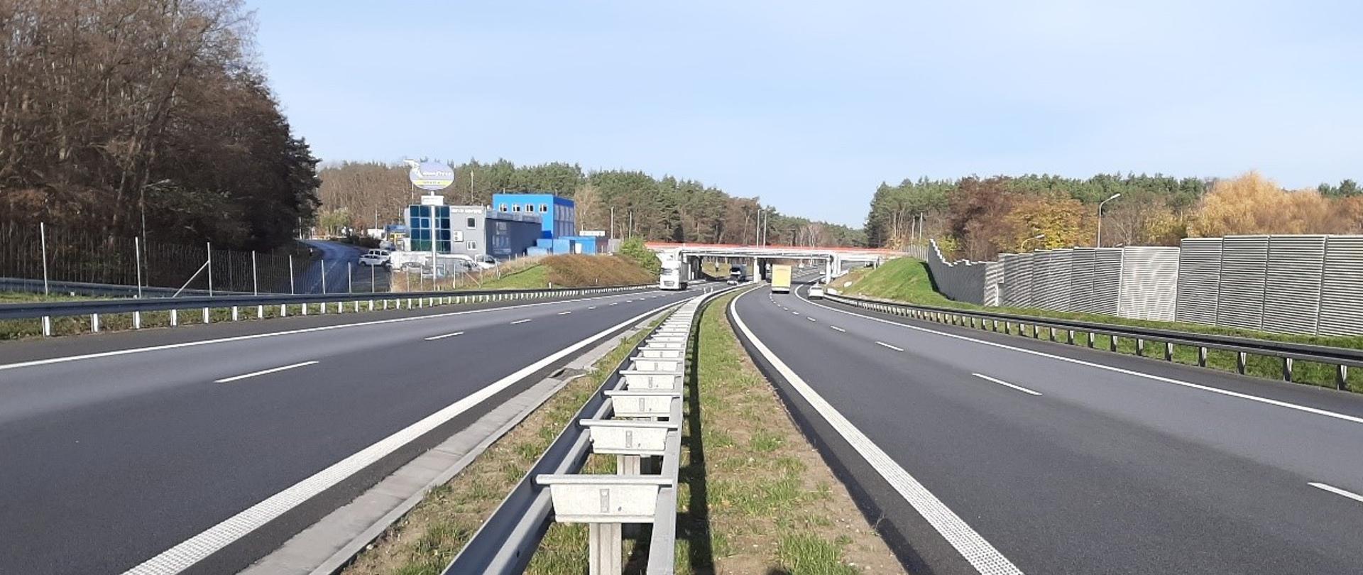 Zdjęcie z poziomu ziemi. Ujęcie w pasie rozdziału między jezdniami. Widoczna bariera ochronna w środku pasa rozdziału. Po prawej stronie ekran akustyczny, po lewej zabudowania, obiekty handlowo-usługowe. W oddali wiadukt nad drogą.
