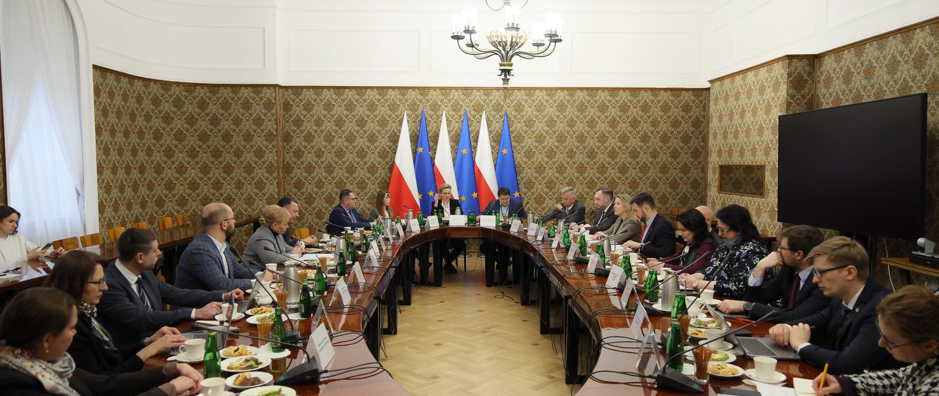 Posiedzenie Komitetu do spraw Pożytku Publicznego w Kancelarii Prezesa Rady Ministrów 18 lutego 2026 roku. Szeroki kadr. Na zdjęciu członkowie Komitetu siedzą przy owalnym stole. W tle przy ścianie są flagi Polski i Unii Europejskiej. Po prawej stronie widoczny jest telewizor, po lewej stronie widać okno.