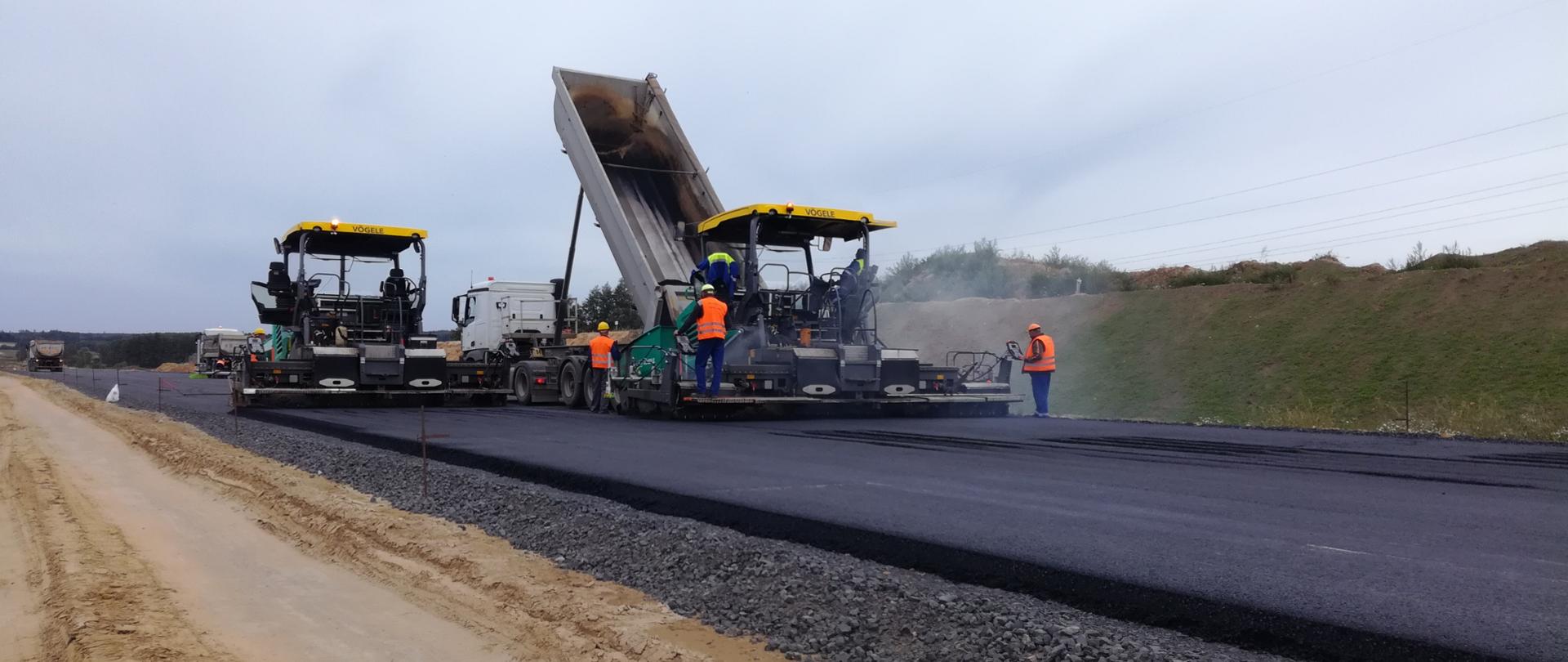 Zdjęcie przedstawia maszyny tzw. rozściełacze podczas pracy, czyli układania masy bitumicznej na nowej drodze. 