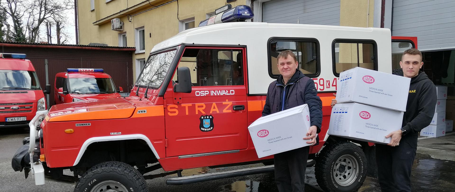 Widok z przodu. Dwaj strażacy OSP stoją na tle samochodu OSP i trzymają w rękach paczki z maseczkami ochronnymi. 