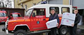Widok z przodu. Dwaj strażacy OSP stoją na tle samochodu OSP i trzymają w rękach paczki z maseczkami ochronnymi. 