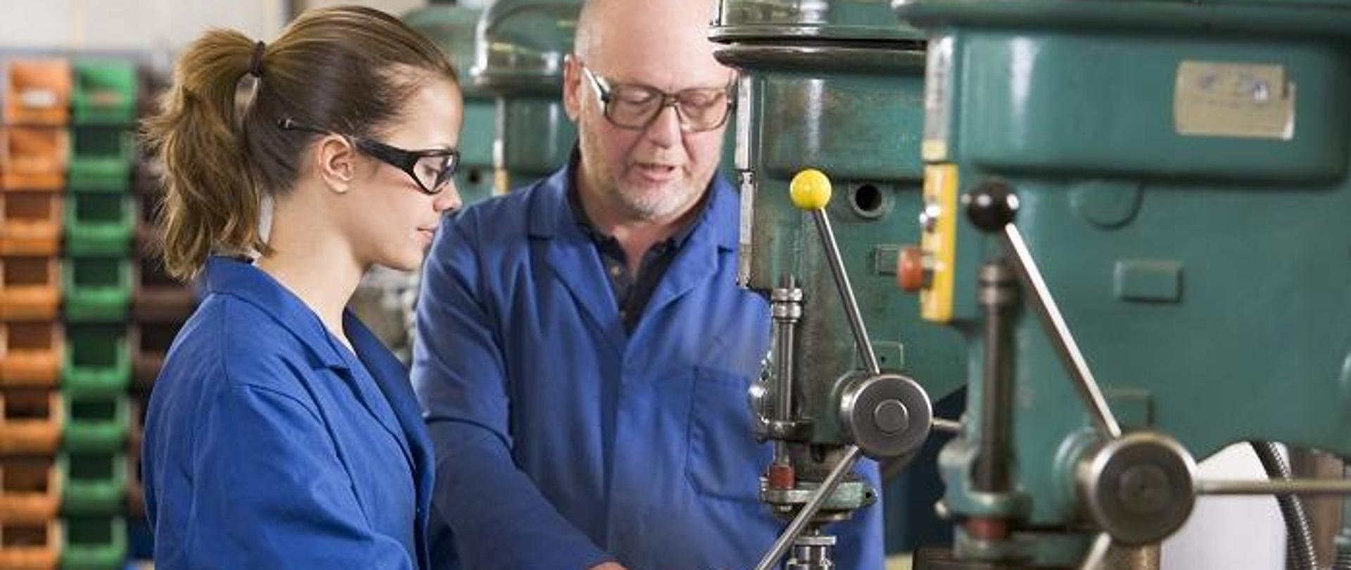 Mistrz i uczennica w warsztacie tokarskim, stoją przy wiertarce kolumnowej