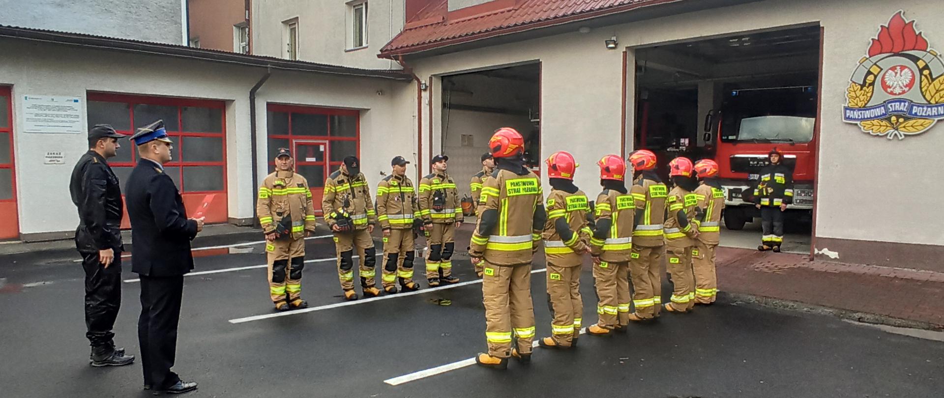 Na zdjęciu widać funkcjonariuszy PSP podczas zmiany służbowej. Zmiana zdająca i przyjmująca stoją na przeciwko siebie. Przed nimi stoi komendant powiatowy wraz z zastępcą dowódcy jednostki. Komendant ubrany jest w mundur galowy i trzyma w ręku akty mianowania. Widać garaże JRG a w nich samochody pożarnicze. Zdjęcie wykonane jest na placu Komendy Powiatowej PSP w Ustrzykach Dolnych.