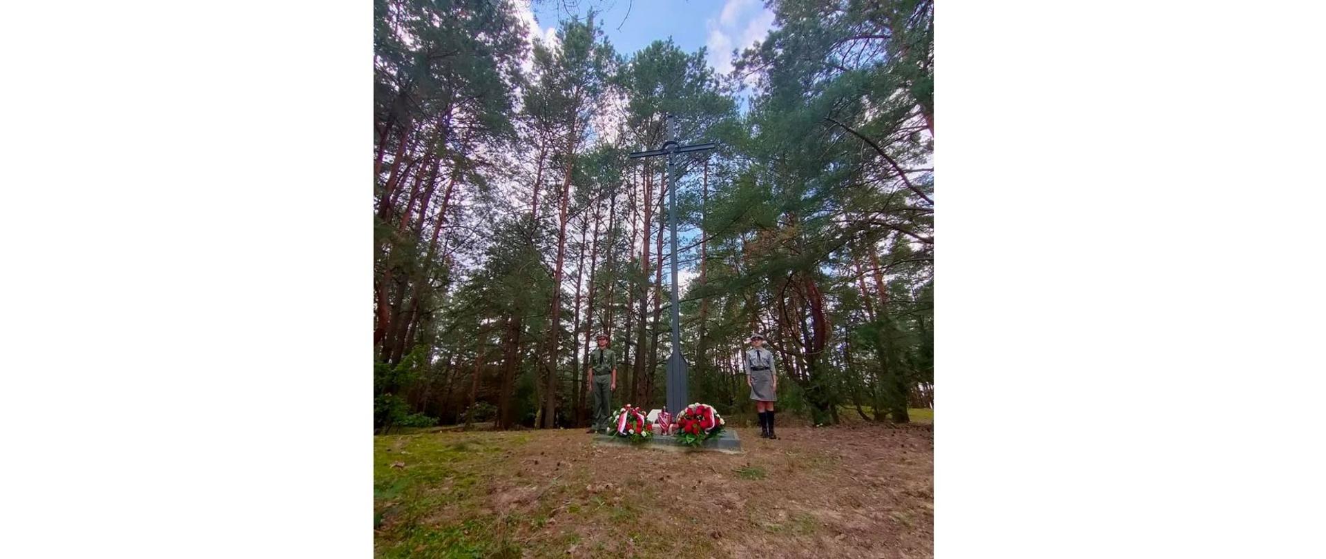 Zdjęcie przedstawia wysoki krzyż usytuowany wśród wysokich drzew, na najwyższym wzniesieniu w okolicy. 