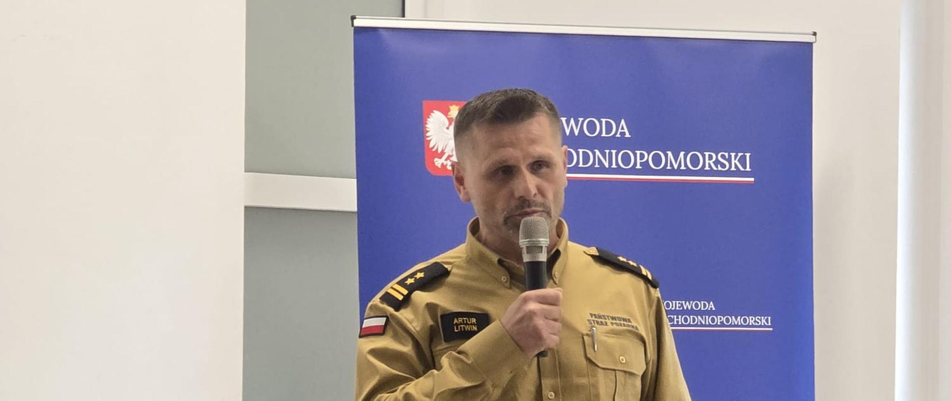 Funkcjonariusz Państwowej Straży Pożarnej przemawia do uczestników spotkania. W tle widoczny baner Wojewody Zachodniopomorskiego.
