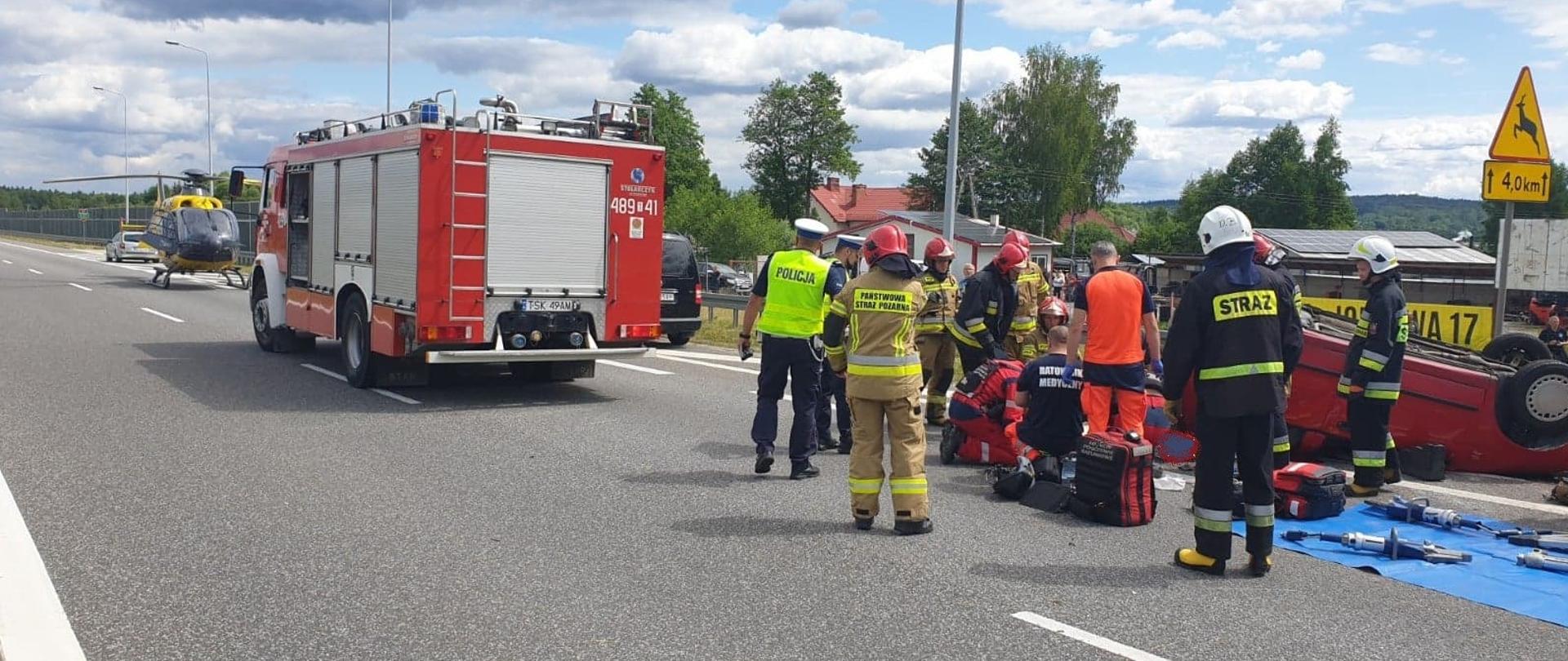 Zdjęcie obrazuje dachowanie samochodu osobowego na trasie S7. Ratownicy medyczni wraz z LPR i strażakami udzielają pomocy medycznej osobie poszkodowanej w zdarzeniu. Obok nich stoją również policjanci. Z prawej strony widać rozłożony sprzęt hydrauliczny na macie sprzętowej. Z lewej natomiast samochód pożarniczy i śmigłowiec LPR.