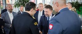 Minister Spraw Wewnętrznych i Administracji podający dłoń Komendantowi Głównemu Państwowej Straży Pożarnej. Wokół nich osoby obserwujące sytuację 