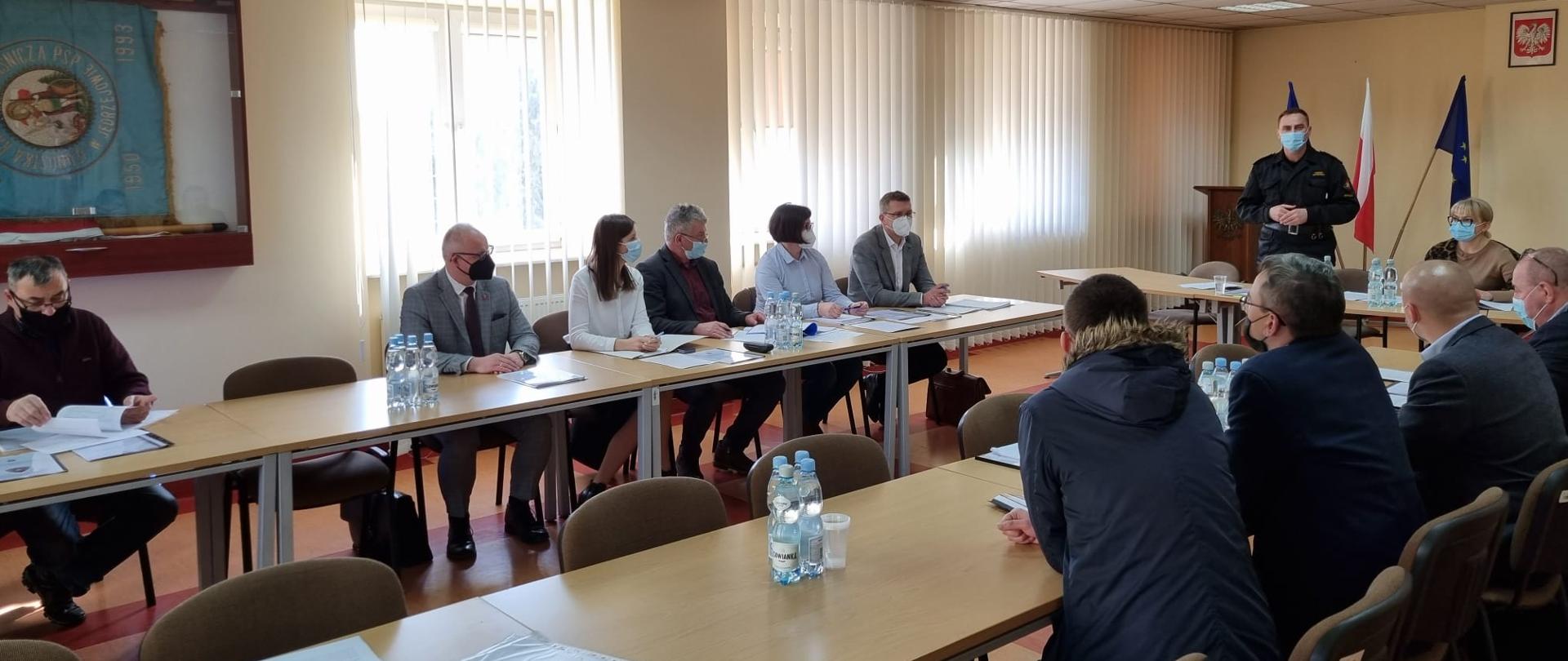 Zdjęcie przedstawia salę konferencyjną KP PSP w Jędrzejowie. Na sali zgromadzeni są przedstawiciele samorządów terytorialnych z terenu powiatu jędrzejowskiego. Przed zaproszonymi gośćmi przy stole na stojąco przemawia zastępca komendanta powiatowego PSP w Jędrzejowie bryg. Tomasz Szymański. Wszyscy uczestnicy mają założone maseczki ochronne zakrywające usta i nos.