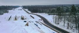Zimowy widok z drona na odcinek drogi w budowie biegnący przez las, z maszynami pracującymi po obu stronach trasy.