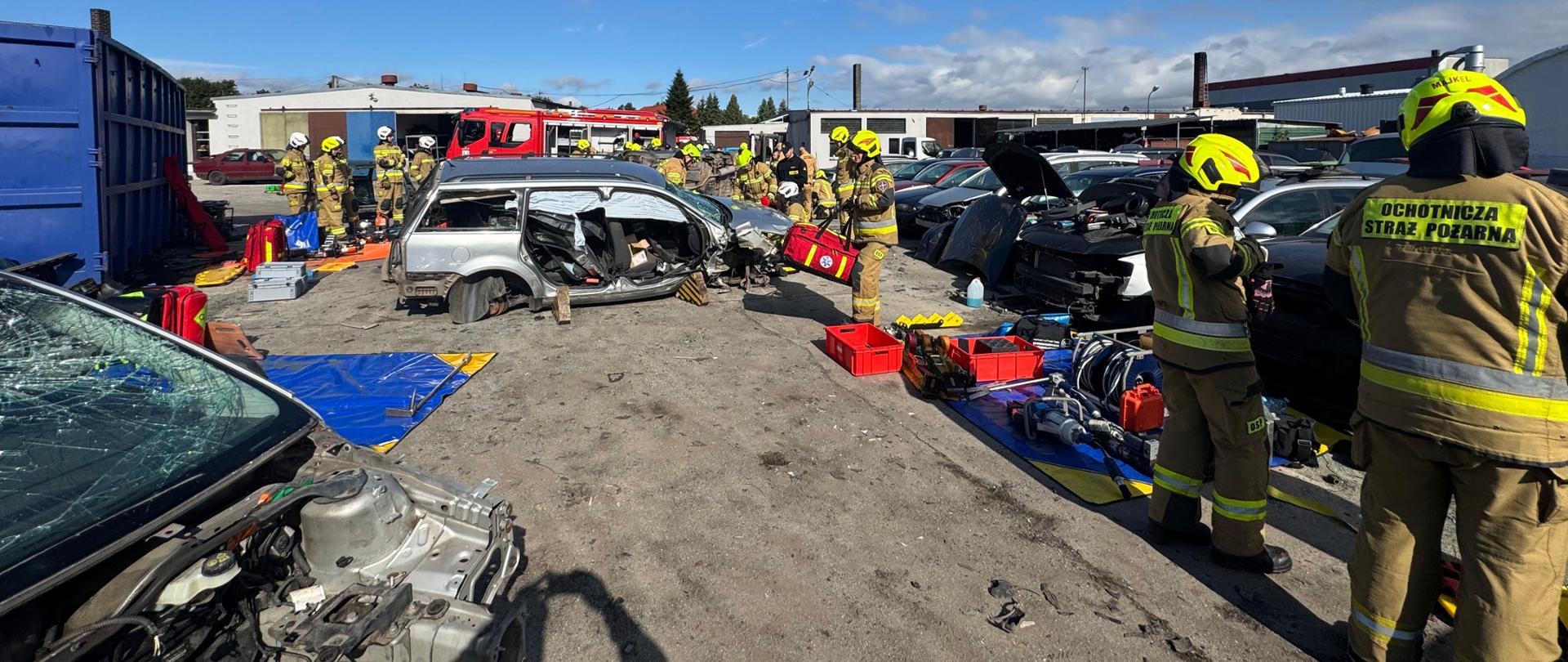 Strażacy w pełnym umundurowaniu uczestniczą w warsztatach z ratownictwa technicznego i medycznego na terenie złomowiska. Na pierwszym planie widoczne są uszkodzone pojazdy, narzędzia hydrauliczne i sprzęt medyczny. Strażacy Ochotniczej Straży Pożarnej (OSP) wykonują symulację akcji ratowniczej po wypadku drogowym. W tle stoją wozy strażackie oraz większa grupa ratowników przygotowujących się do działania. Pogoda słoneczna, niebo bezchmurne.