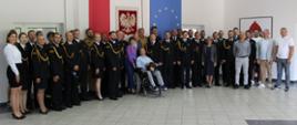 grupa osób stojąca na tle godła RP flagi Polski oraz flagi Unii Europejskiej część obrona mundury a druga część pobrania cywilna z przodu przednimi wózek na wózku inwalidzkim siedzi osoba ubrana po cywilnemu która na kolanach trzyma kolorowy hełm strażacki