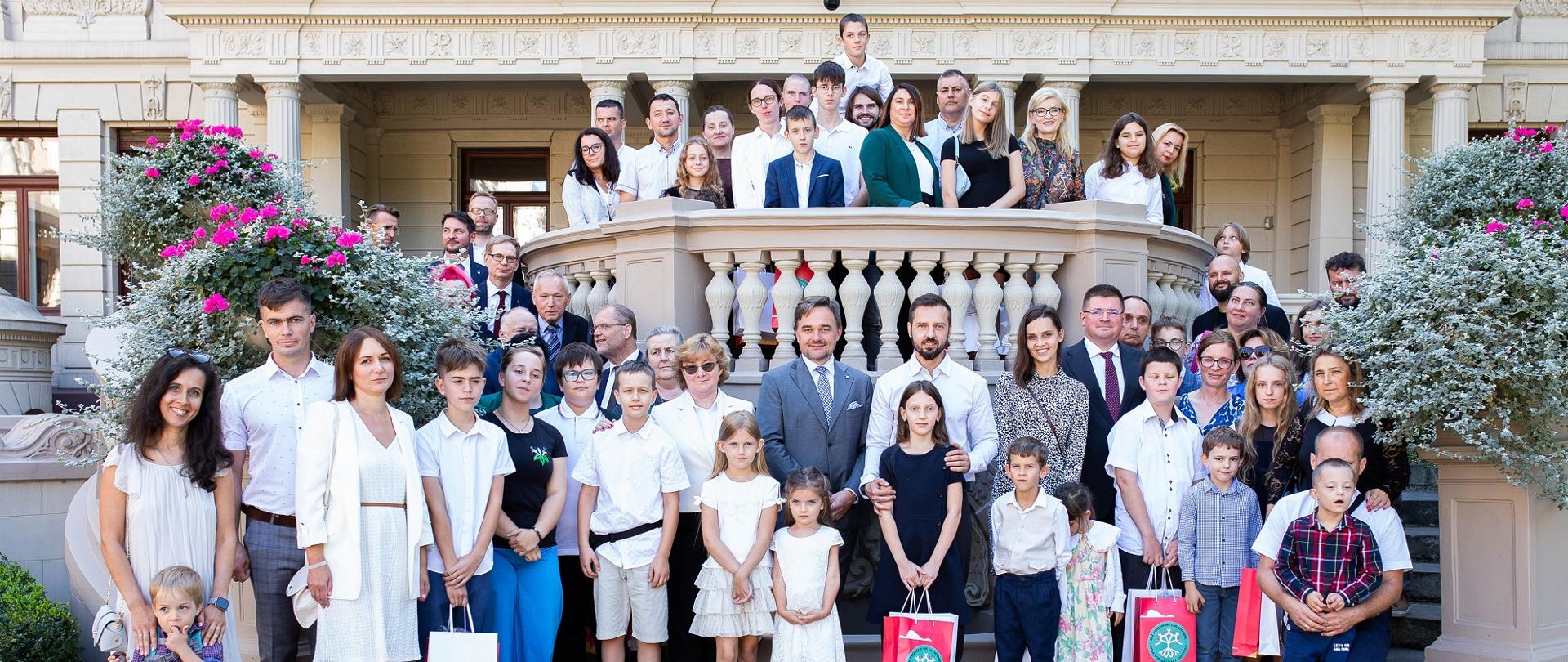 Na dworze przed budynkiem z kolumnami stoi duża grupa ludzi. po bokach krzaki z fioletowymi kwiatkami.