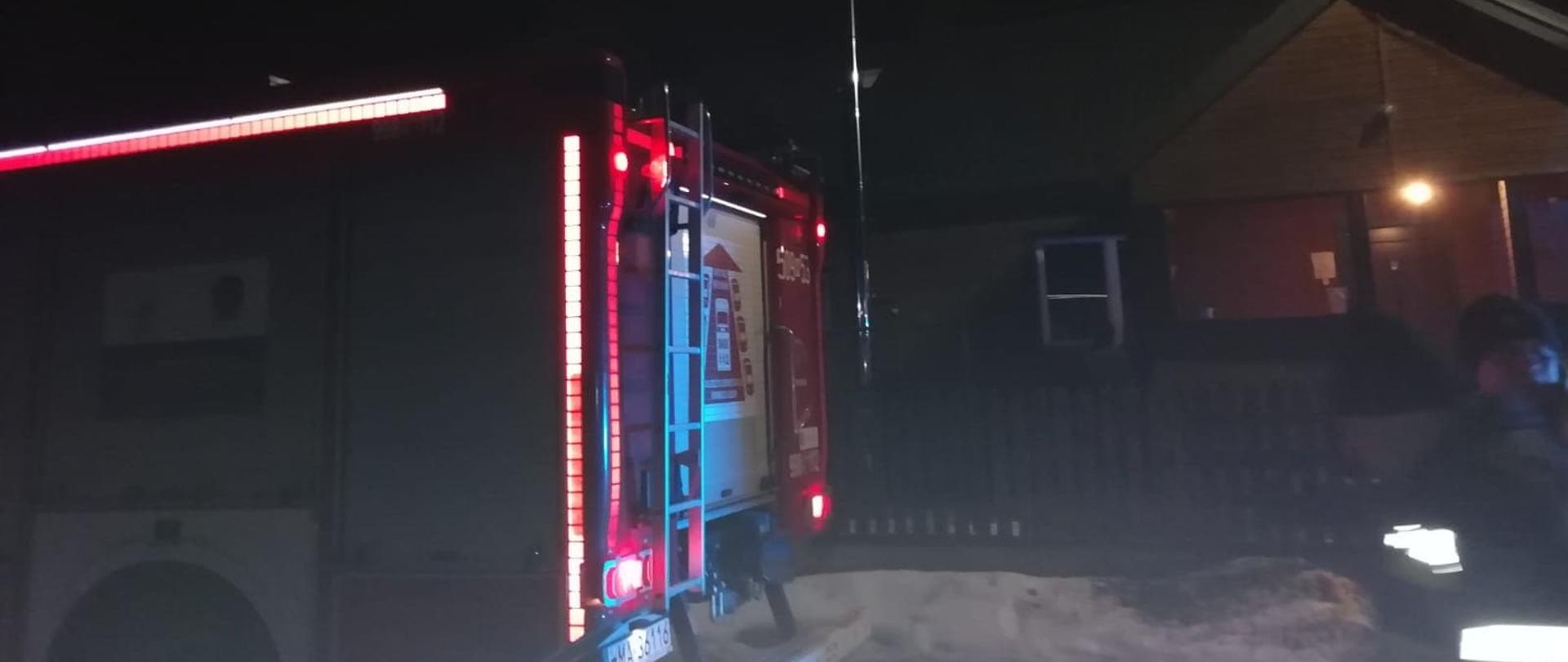 Samochód strażacki stoi zaparkowany przed drewnianym domem jednorodzinnym . Pora nocna.