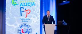 Przy mównicy stoi minister Czarnek i mówi do mikrofonu, za nim na ścianie napisy Fundacja wstawaj Alicja, Fizjoterapia Polska i Akademia Wincentego Pola.
