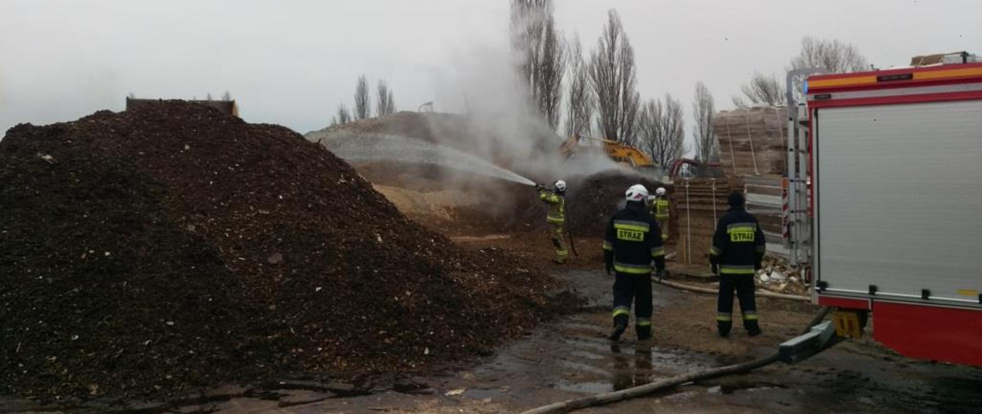 Na zdjęciu widać hałdy odpadów poprodukcyjnych zakładu stolarskiego. Z hałd wydobywają się białe obłoki dymu i pary wodnej. Po prawej stronie przy jednej z hałd widać samochód strażacki w kolorze czerwonym ze srebrnymi elementami zabudowy. Przy pojeździe stoi dwóch strażaków. W oddali widać kolejnych dwóch strażaków polewających palącą się hałdę wodą. Za nimi znajduje się koparka. 