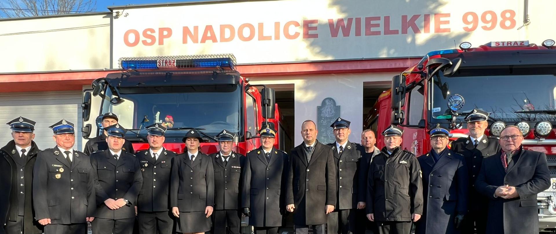 Grupa strażaków oraz osoby cywilne pozują do zdjęcia , w tle 2 samochody bojowe OSP oraz strażnica