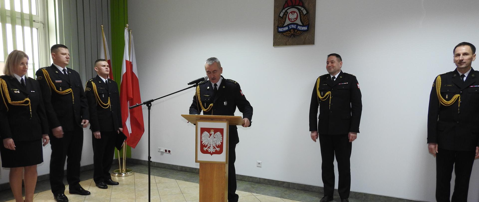 Na pierwszym planie widać zdającego obowiązki komendanta powiatowego Państwowej Straży Pożarnej w Tomaszowie Mazowieckim st. bryg. Krzysztofa Iskierkę, który stoi przed mównicą i zabiera głos do mikrofonu. Po prawej stronie stoi z-ca łódzkiego komendanta wojewódzkiego PSP w Łodzi - st. bryg. Sebastian Kościsty oraz przejmujący obowiązków komendanta PSP w Tomaszowie Mazowieckim - st. bryg. Marcin Dulas. Po lewej stronie stoi trzech funkcjonariuszy. Ma mównicy zawieszone jest godło polski. W tle na ścianie zawieszone jest logo PSP. Za funkcjonariuszami po lewej stronie stoją flagi polski. Strażacy ubrani są w czarne mundury ze złotym sznurem galowym.