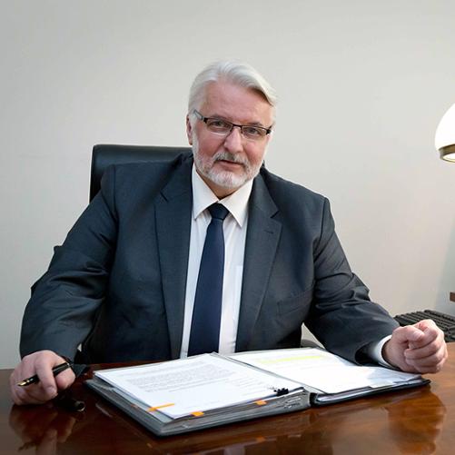 Witold Waszczykowski, kolorowy portret mężczyzny w okularach siedzącego przy biurku, siwe włosy, broda, grafitowa marynarka, granatowy krawat, w prawej słoni pióro, na biurku segregator z dokumentami.