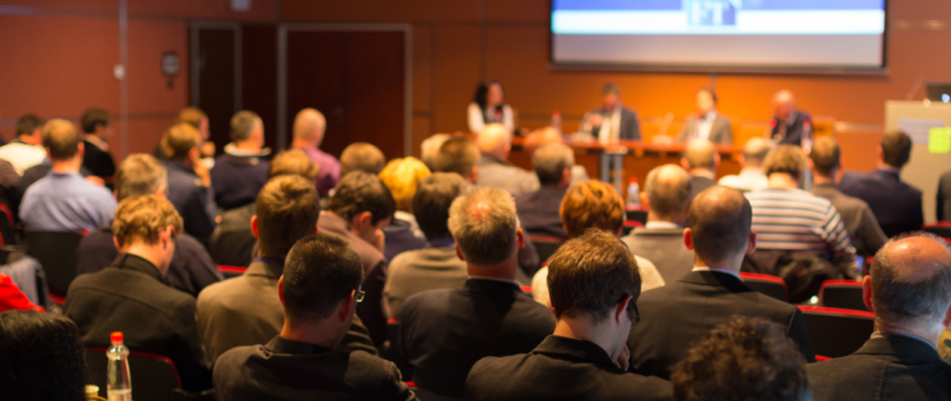 Panel pięciu prelegentów przy stole, publiczność siedzi naprzeciw, w tle ekran z prezentacją.