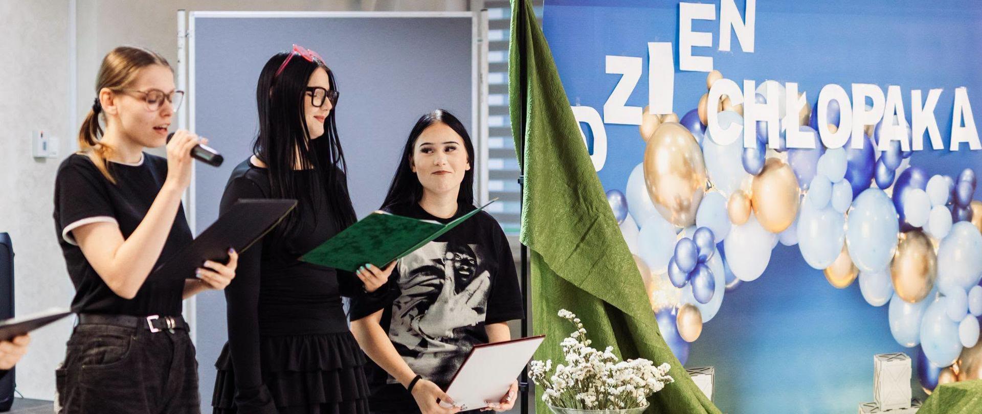 Zdjęcie przedstawia fragment szkolnej uroczystości z okazji Dnia Chłopaka. Po lewej stronie trzy osoby stoją na scenie lub w jej pobliżu. Jedna z nich przemawia przez mikrofon, trzymając czarną teczkę, druga odczytuje informacje z zielonej teczki, a trzecia trzyma dokument lub dyplom. Po prawej stronie znajduje się okolicznościowa dekoracja z dużym napisem „Dzień Chłopaka”, kompozycją balonów w kolorach białym, niebieskim i złotym oraz zieloną tkaniną ułożoną w dekoracyjne fałdy. Całość uzupełniają szklane wazony z białymi kwiatami.