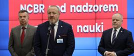 Na tle wbudowanego w ścianę wielkiego ekranu z napisem NCBR pod nadzorem Ministra Nauki stoi minister Wieczorek, obok niego mężczyzna w garniturze który mówi do mikrofonu na stojaku, z tyłu stoi wiceminister Gzik.