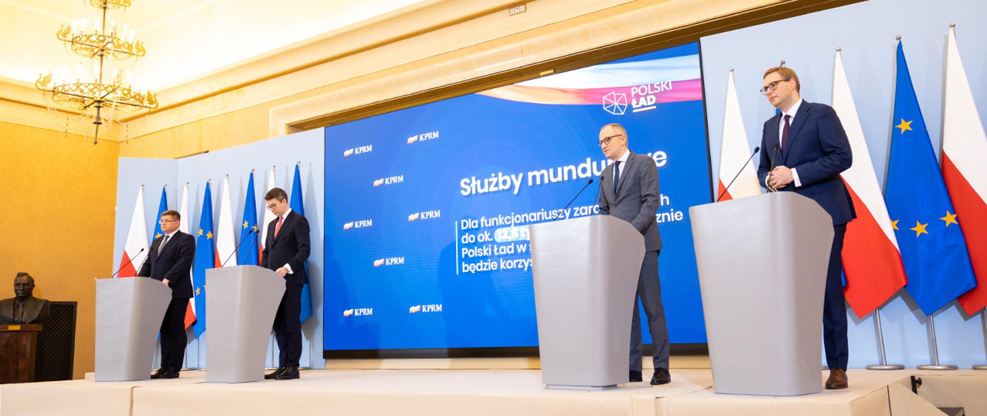 Na zdjęciu widać uczestników konferencji prasowej stojących za mównicami, m.in wiceministra Błażeja Pobożego. W tle widać flagi Polski i UE i ekran z wyświetloną prezentacją.