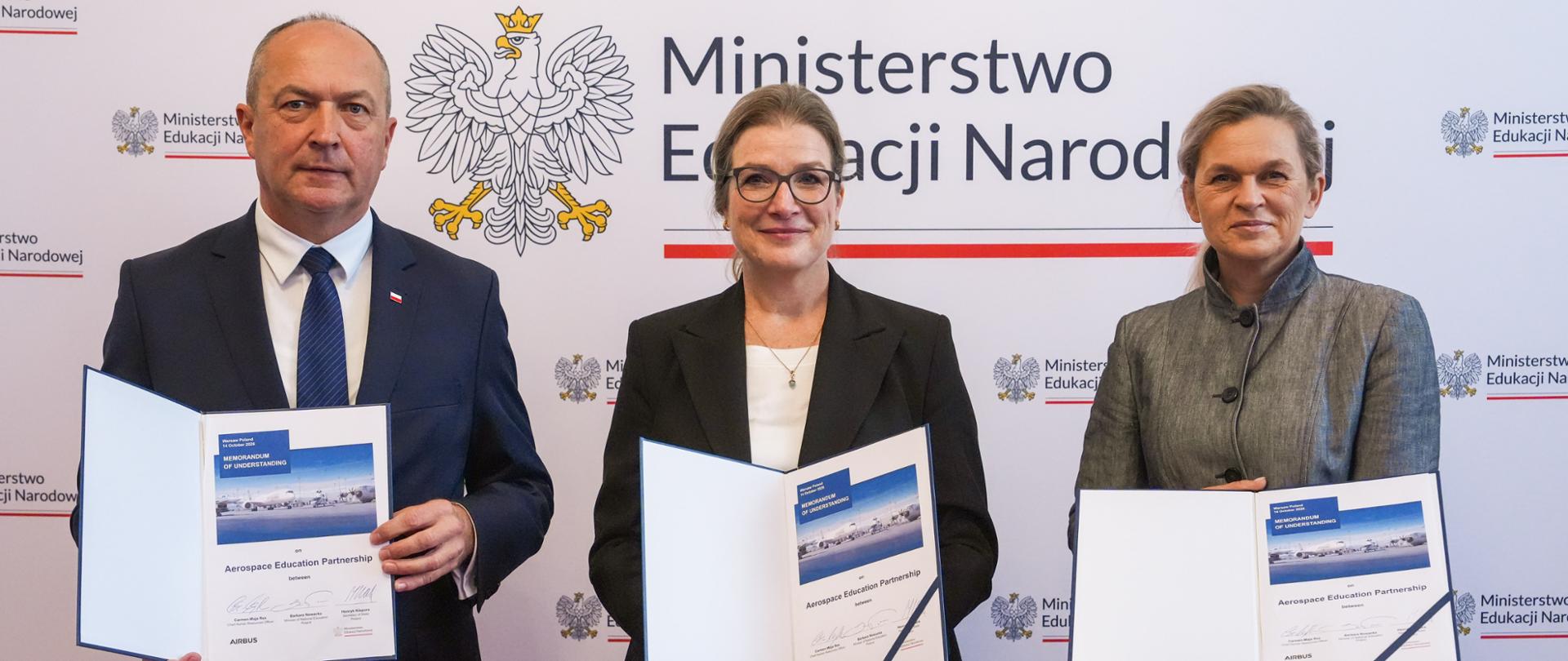 Trzy osoby (mężczyzna i dwie kobiety) stojące na tle godła Polski oraz napisu „Ministerstwo Edukacji Narodowej”, prezentujące otwarte dokumenty z podpisami