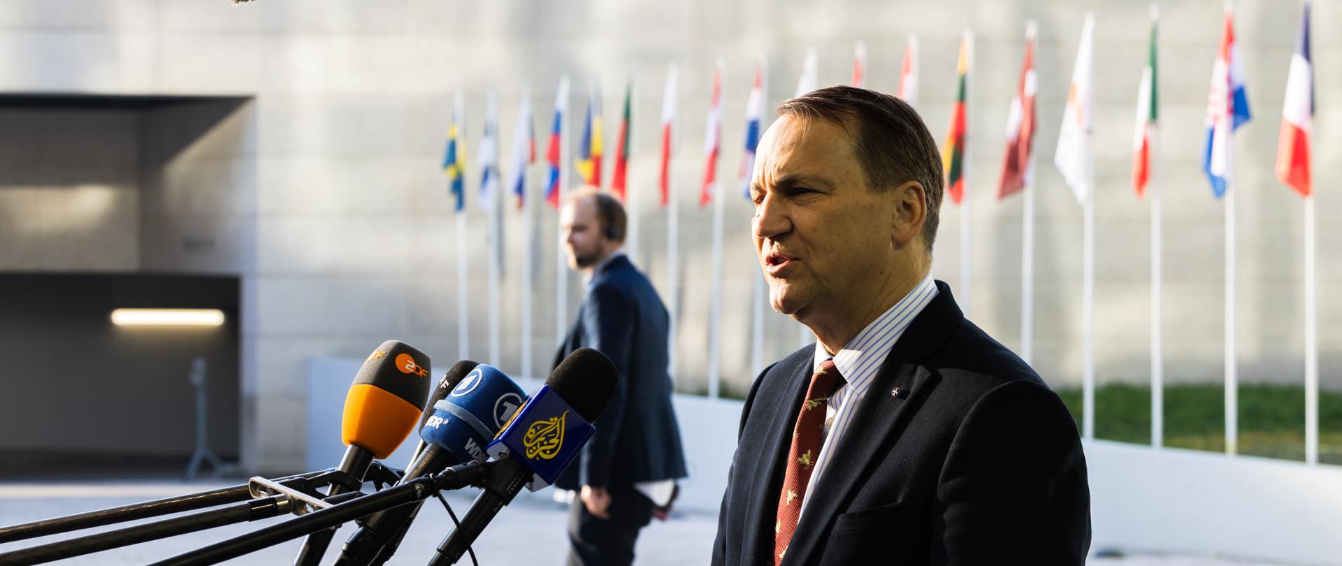 Mężczyzna stojący na tle flag mówi do mikrofonów.