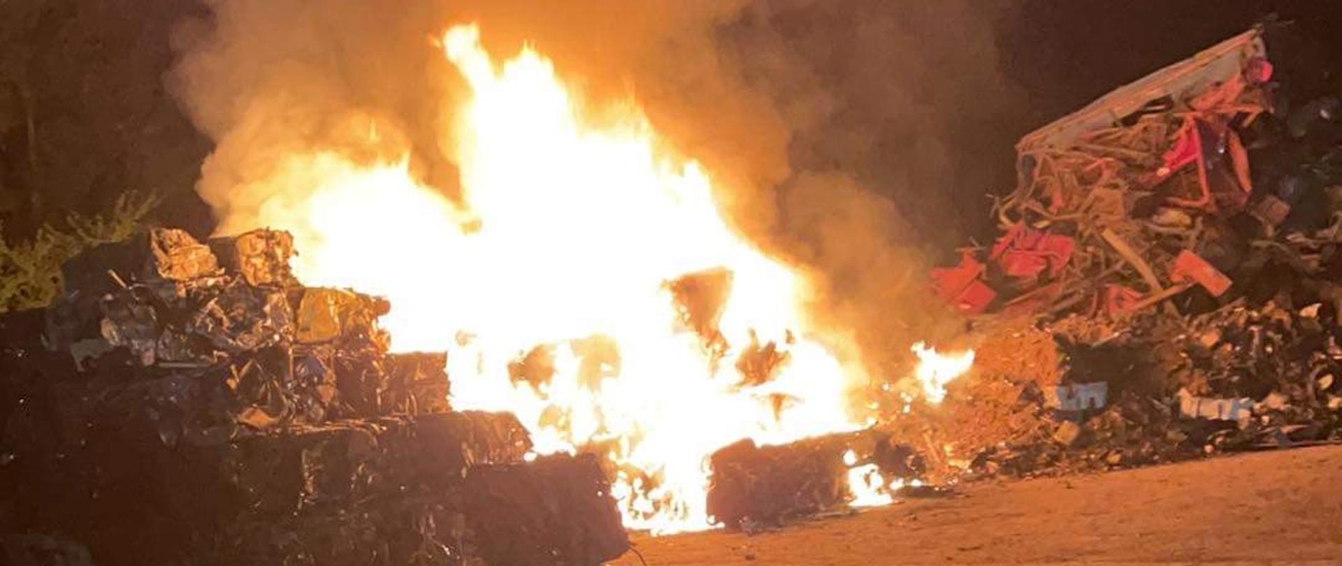 Zdjęcie przedstawia palące się sprasowane kostki samochodów osobowych na złomowisku. Pora nocna. 