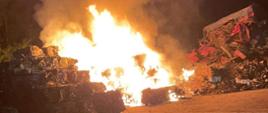 Zdjęcie przedstawia palące się sprasowane kostki samochodów osobowych na złomowisku. Pora nocna. 