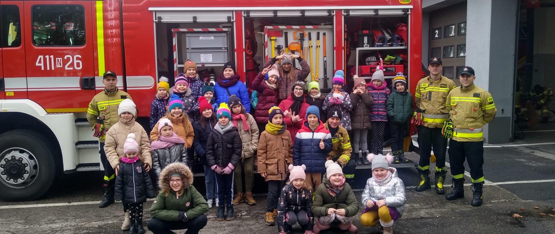 Zdjęcie zrobione na placu przed remiza państwowej straży pożarnej. Na tle dużego czerwonego samochodu strażackiego stoi duża grupa kolorowo ubranych dzieci. Z prawej strony otwarte bramy garaży i dwóch strażaków.