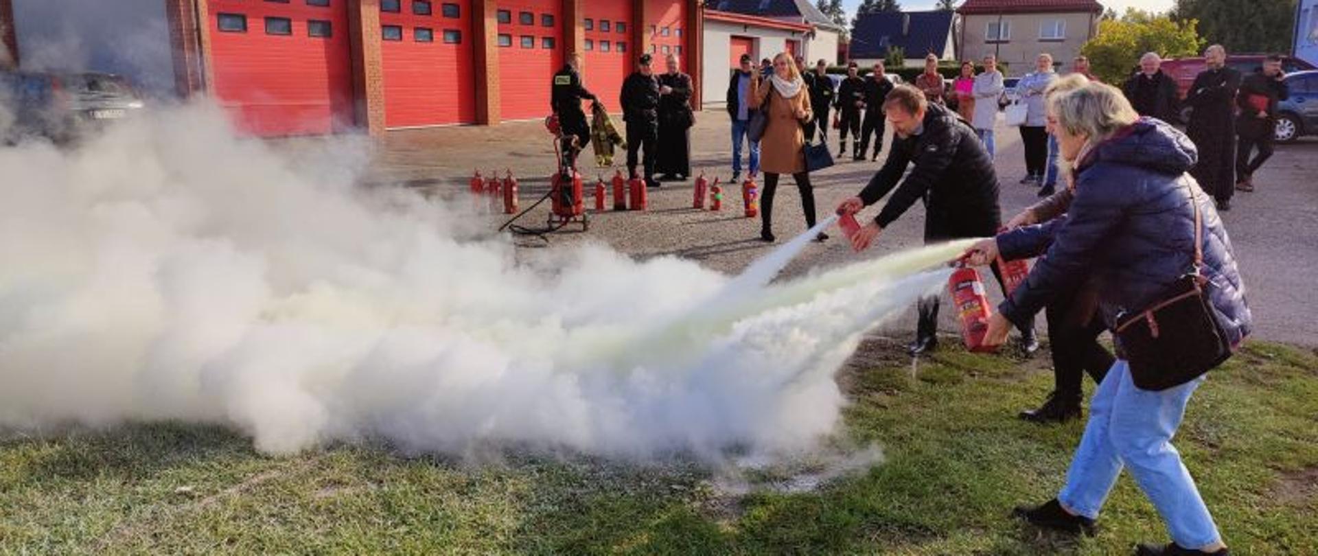 Zdjęcie wykonane na zewnątrz budynku. Trzy osoby gaszą palącą się benzynę. Każda z osób ma swoją gaśnicę. W tle reszta grupy obserwująca moment gaszenia.