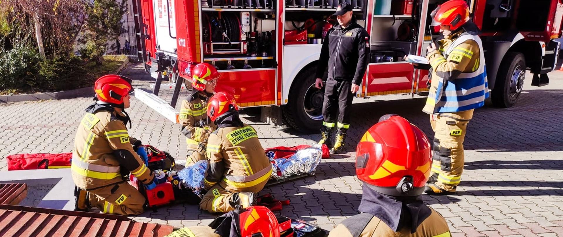 Na zdjęciu widać samochód pożarniczy Man w którym otwarte są skrytki. Przy samochodzie tym stoi strażak w ubraniu koszarowym który patrzy w kierunku wykonujących założenie strażaków. Strażacy ubrani w ubrania specjalne znajdują się przy dwóch poszkodowanych. Przy jednym poszkodowanym którym jest manekin znajduje się trzech strażaków przy drugim poszkodowanym jest dóch strażaków. Wszyscy Ci strażacy klęczą z wyjątkiem strażaka w koszarówce i dowódcy który ma na sobie kamizelkę w kolorze niebieskim i stoi nad wszystkimi.