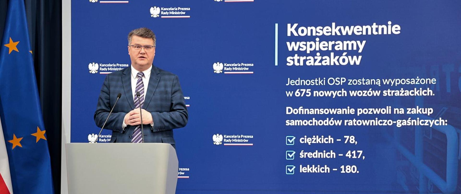 Wiceminister Maciej Wąsik przemawia przy mównicy w trakcie konferencji wspieramy strażaków w tle opis Konsekwentnie wspieramy strażaków, Jednostki OSP zostaną wyposażone w 675 nowych wozów strażackich. Dofinansowanie pozwoli na zakup samochodów ratowniczo-gaśniczych: ciężkich -78, średnich-417 i lekkich -180.