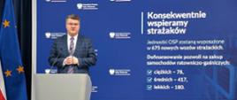 Wiceminister Maciej Wąsik przemawia przy mównicy w trakcie konferencji wspieramy strażaków w tle opis Konsekwentnie wspieramy strażaków, Jednostki OSP zostaną wyposażone w 675 nowych wozów strażackich. Dofinansowanie pozwoli na zakup samochodów ratowniczo-gaśniczych: ciężkich -78, średnich-417 i lekkich -180.