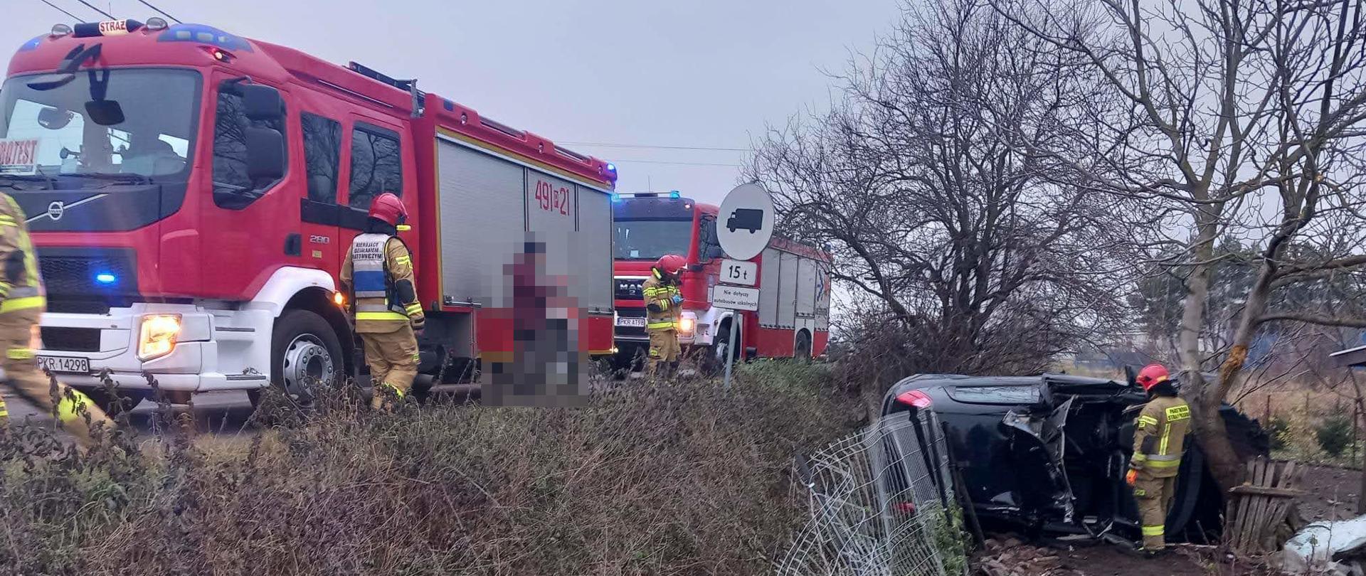 Samochód osobowy leży na boku poza jezdnią. W pojeździe usunięto fragment dachu. Obok zdeformowanego pojazdu stoi strażak. Na jezdni wzdłuż miejsca wypadku stoją wozy pożarnicze oraz pozostała część obsady. Na pierwszym planie fragment uszkodzonego płotu, który staranował samochód