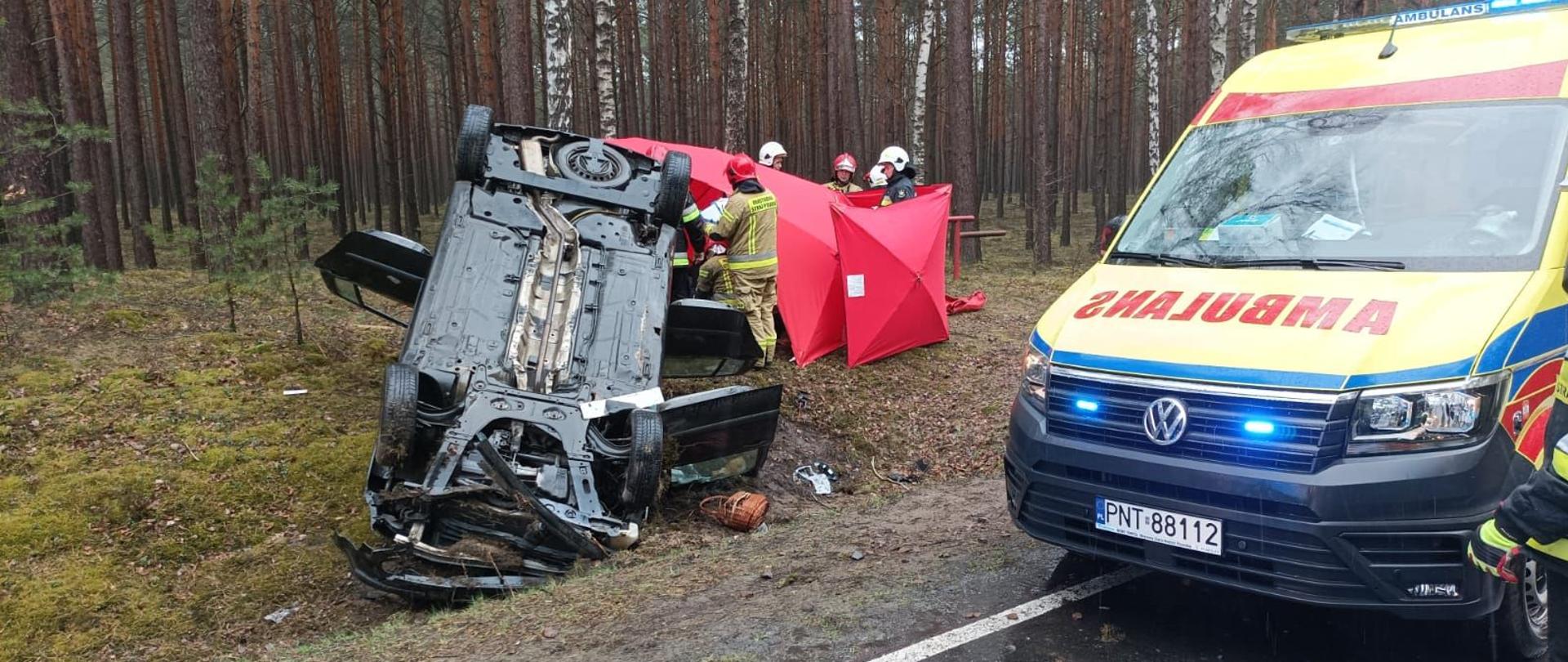 Samochód osobowy leżący na dachu, obok karetka pogotowia ratunkowego, w tle strażacy za parawanami udzielający pomocy.