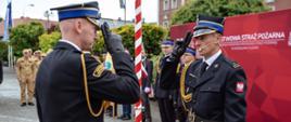 Dwaj oficerowie Państwowej Straży Pożarnej w mundurach galowych salutują sobie nawzajem podczas uroczystego apelu przy biało-czerwonym maszcie flagowym.