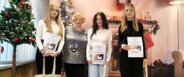 Laureatki konkursu i organizatorka pozują do zdjęcia obok choinki i dekoracyjnego kominka. Każda z nagrodzonych osób trzyma dyplom oraz materiałową torbę z nadrukiem budynku szkoły. Obok nich leżą zapakowane prezenty.