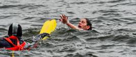 Strażak ubrany w piankę płynie do osoby poszkodowanej znajdującej się w wodzie. Strażak posiada czerwoną uprząż zabezpieczającą. Strażak podaje kobiecie znajdującej się w wodzie pas ratowniczy koloru żółtego.