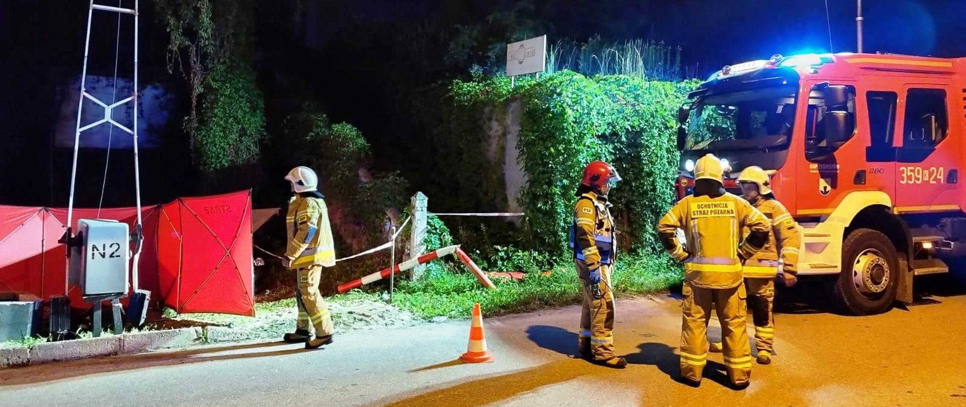 Strażacy i samochód pożarniczy na drodze. Obok rogatki kolejowej rozłożony parawan.