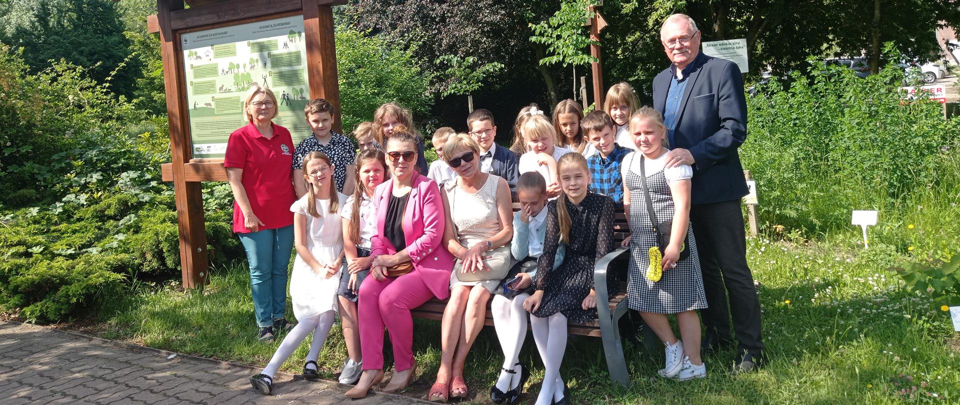 Na ławce wśród zieleni siedzi grupa kilkunastu dziewczynek i chłopców. Z tyłu grupy stoi mężczyzna i kobieta. Wokół rosną drzewa oraz stoją tablice edukacyjne.