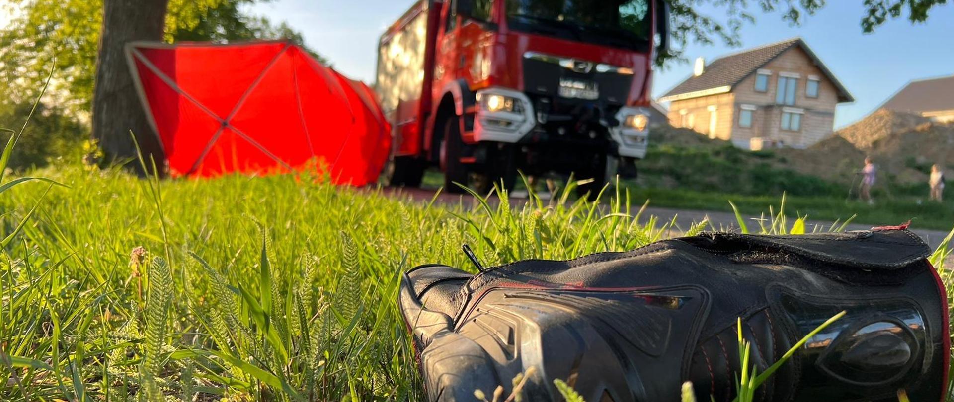 But motocyklisty leżący na trawie poza jezdnią. W tle stoi wóz strażacki na drodze, obok niego rozłożony jest parawan. 