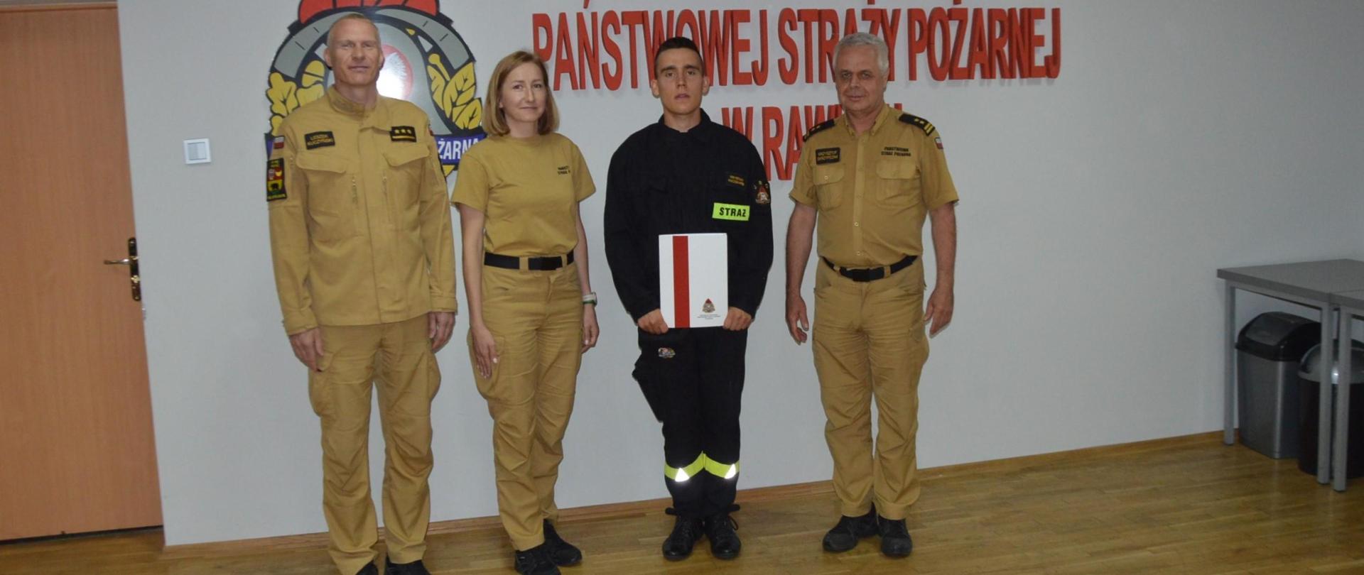 Świetlica Komendy Powiatowej PSP w Rawiczu. Na tle ściany, na której znajduje się czerwony napis: Komenda Powiatowa Państwowej Straży Pożarnej w Rawiczu stoi awansowany strażak w towarzystwie zastępcy komendanta powiatowego PSP oraz zastępcy dowódcy JRG i funkcjonariuszki d/s kadrowych. 