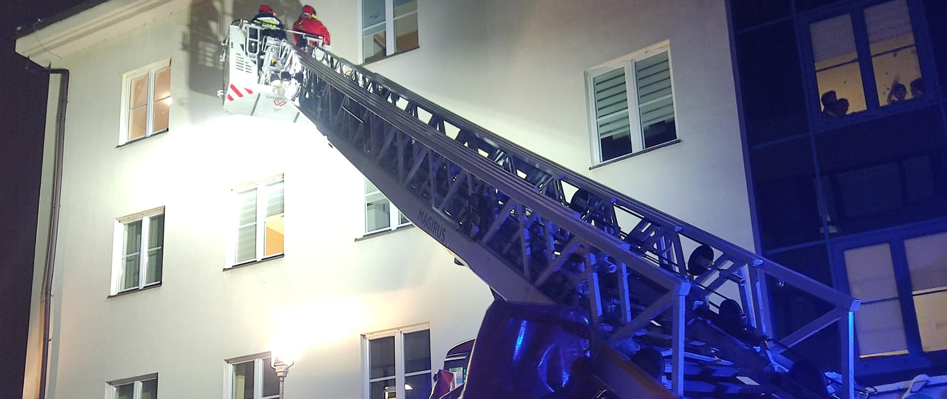 Drabina mechaniczna rozstawiona przed budynkiem szpitalnym oddziału Św. Mikołaja z drabina wysuniętą do 3 piętra. W koszu znajduje się Święty Mikołaj oraz strażak. W oknie widać dzieci.