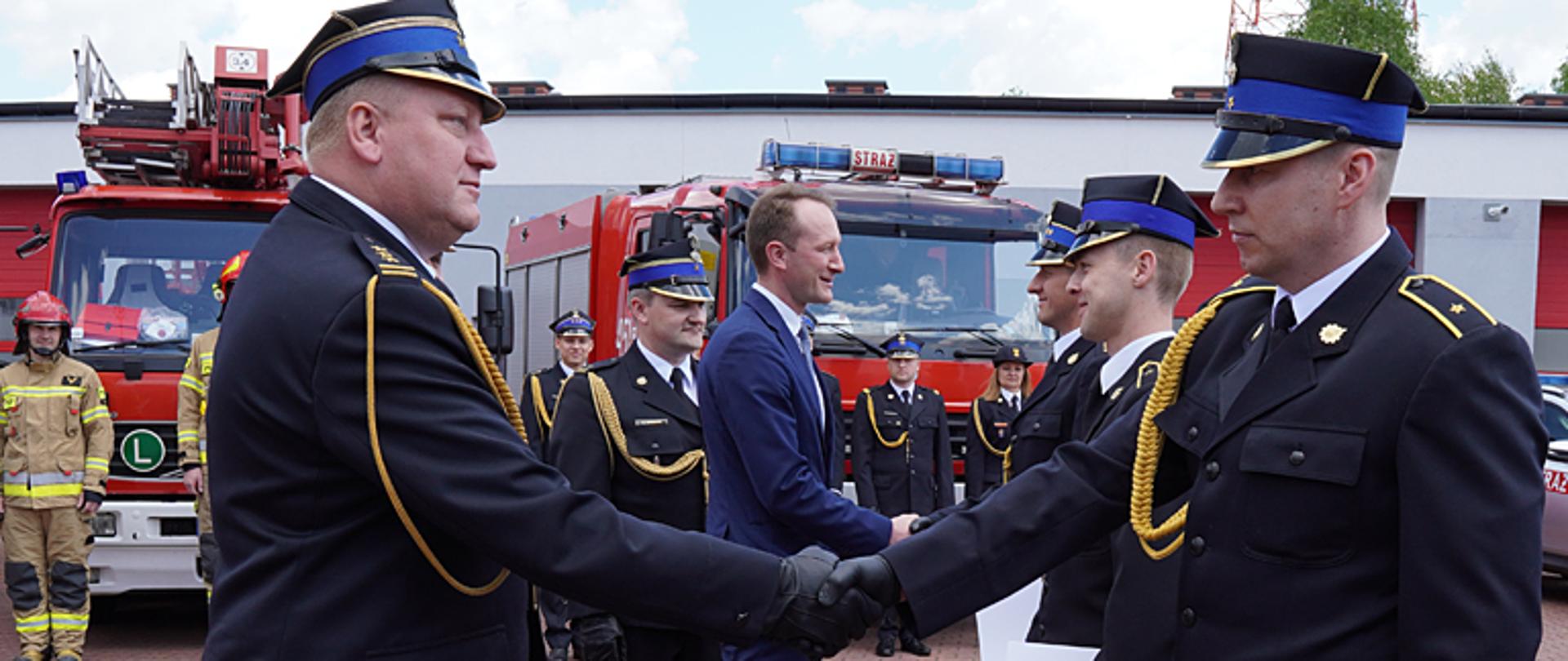 Zdjęcie przedstawia komendanta wojewódzkiego st. bryg. Grzegorza Janowskiego gratulującego awansu strażakowi, podają sobie prawe dłonie w uścisku, na drugim planie wicestarosta łowicki Piotr Malczyk gratuluje awansu strażakowi podając mu prawą dłoń. W tle zastępca komendanta powiatowego mł. bryg. Rafał Garbarz i pojazdy straży pożarnej, widać budynek garażowy a na jego tle strażaków stojących w szeregu. Funkcjonariusze ubrani są w umundurowanie galowe ze sznurem, na głowie mają rogatywkę oraz czarne rękawiczki.