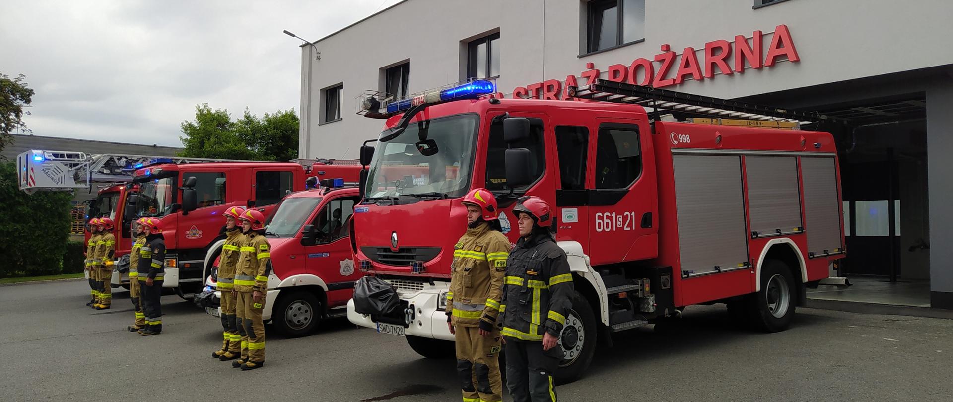 Na zdjęciu strażacy na tle samochodów pożarniczych oraz remizy