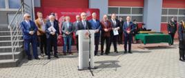 Zdjęcie przedstawia Zastępcę Prezydenta Miasta Konina, który stoi przy mównicy na placu przed budynkiem komendy podczas uroczystych obchodów dnia strażaka. Z tyłu stoi wicestarosta koniński oraz wójtowi i burmistrzowie powiatu konińskiego