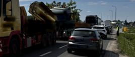 Na pierwszym tle (od lewej): zatrzymane do kontroli 40-tonowe zestawy ciężarowe na odcinku drogi powiatowej, gdzie obowiązuje zakaz tonażowy dla pojazdów o dopuszczalnej masie całkowitej powyżej 15 ton. Przeciwległym pasem ruchu poruszają się samochody osobowe. W tle: ruchem kołowym kieruje ręcznie funkcjonariusz mazowieckiej Inspekcji Transportu Drogowego.