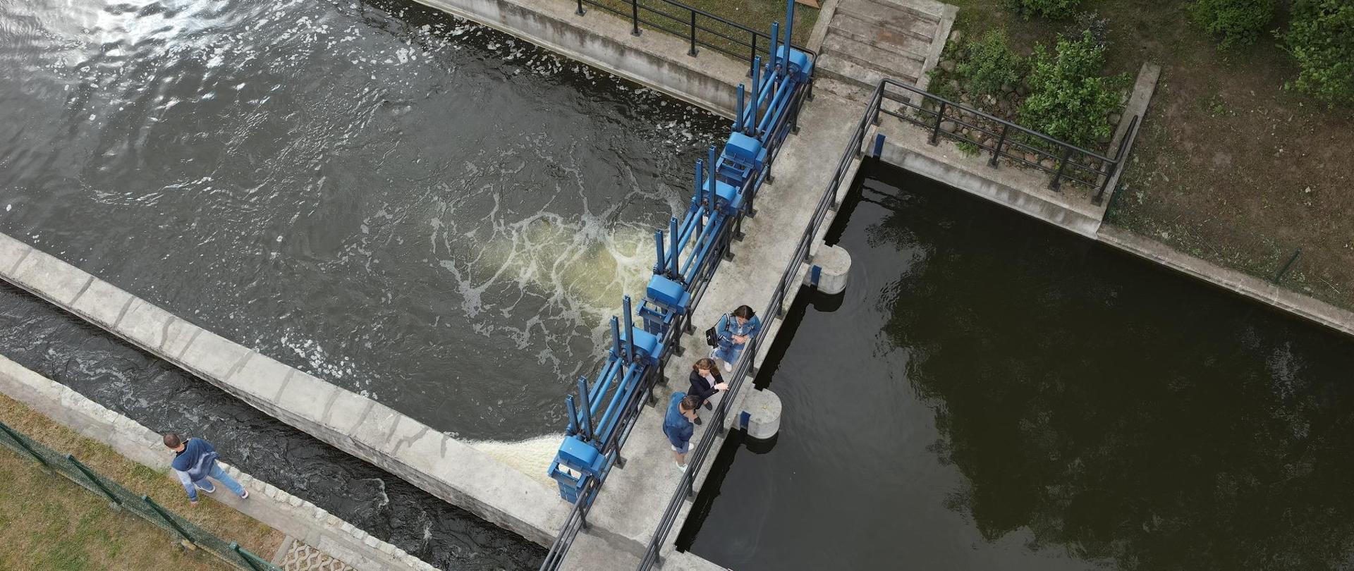 Jaz na rzece Panna na wypływie z Jeziora Mogileńskiego. Ujęcie z lotu ptaka. Na jazie znajdują się turyści i pracownicy Wód Polskich.